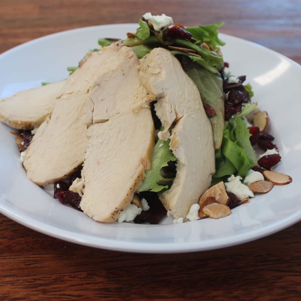 A white plate holds a salad with sliced chicken, cranberries, almonds, and feta cheese.