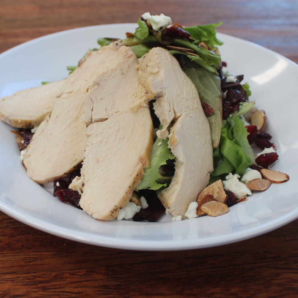 A white plate holds a salad with sliced chicken, cranberries, almonds, and feta cheese.