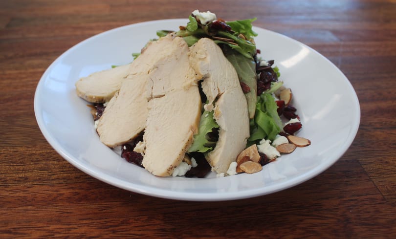A white plate holds a salad with sliced chicken, cranberries, almonds, and feta cheese.