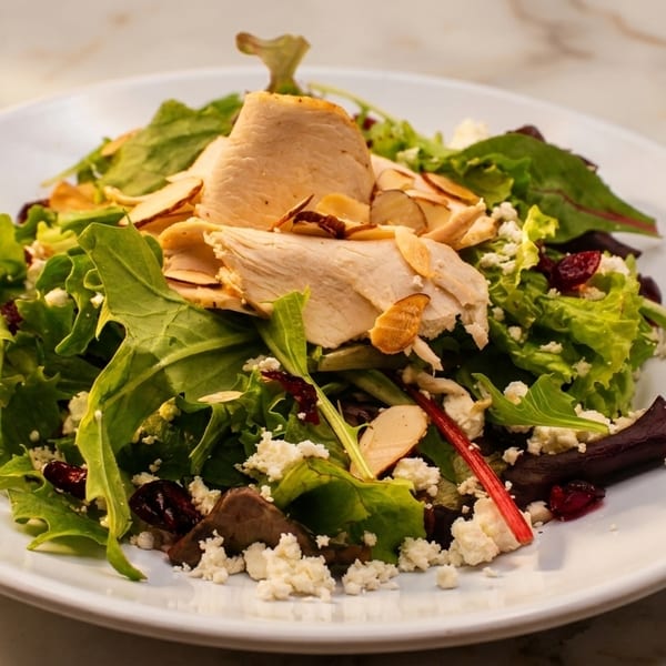 A vibrant salad with sliced chicken, mixed greens, cranberries, feta cheese, and almonds on a white plate.